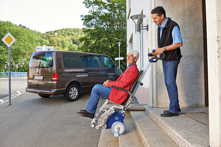 Das Bild zeigt einen Mann mittleren Alters, der einen älteren Mann, der in einem Treppensteiger mit integrierter Sitzeinheit sitzt, die Außentreppe eines Hauses hinunterbefördert. Im Hintergrund steht ein Fahrzeug für den Personentransport / Flughafentransport.